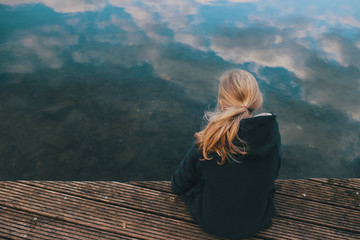 young girl with long blonde hair is sitting on a wooden stage looking in clear shallow water with reflecting cloudy sky in a thoughtful pose - coming of age, sadness and sorrow concept - obrazy, fototapety, plakaty