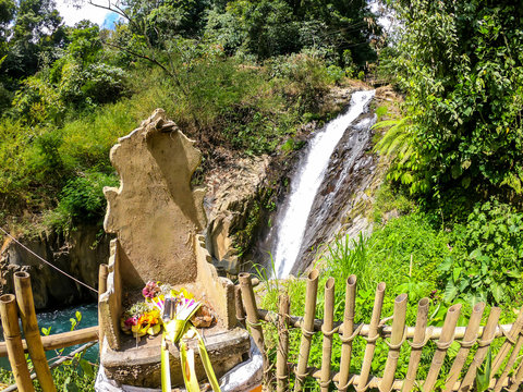 An offering altar with on Gitgit Twin Waterfall, Bali, Indonesia. The waterfall is surrounded by lush green flora, in a dense jungle. The altar resembles a throne, few offerings on it.