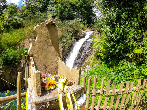 An offering altar with on Gitgit Twin Waterfall, Bali, Indonesia. The waterfall is surrounded by lush green flora, in a dense jungle. The altar resembles a throne, few offerings on it.