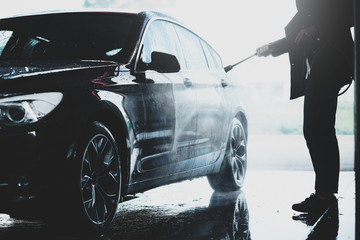 Silhouette of a man washing his black car outdoors in a carwash station, using water jet with soap and high pressured water stream