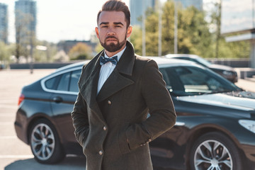 Cool and elegant businessman standing near his car outdoors posing for the photo, wearing white...