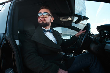 Confident businessman sitting inside of his car, holding a rudder, wearing white shirt, sunglasses, bow tie and dark coat, looking cool and successful