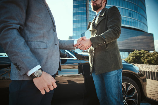 Photo of a two men shaking hands in front of a black car standing outdoors, wearing stylish clothes. Close up shoot