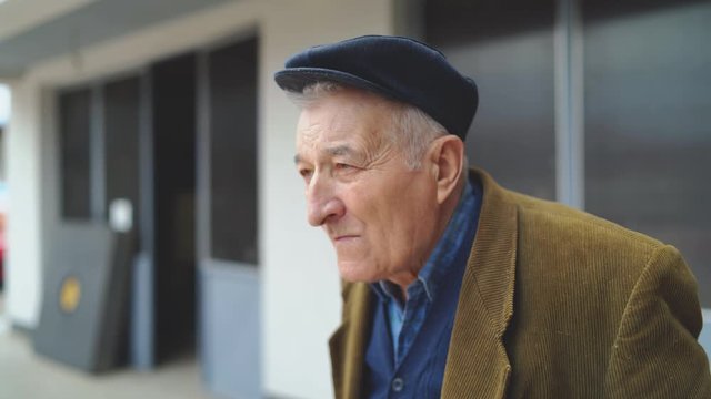 Portrait of senior man pensioner  looking around while standing alone outdoor wearing jacket and hat cap in front of house 