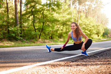 Young woman warming up at forest road before jogging. Attractive girl stretching for running exercises. Beautiful curly sporty girl doing fitness outdoor