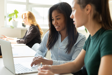 Smiling millennial multiracial female employees cooperate working together on laptop in shared office, diverse women coworkers busy collaborating discuss ideas brainstorm at business meeting