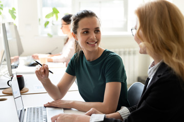 Smiling millennial female employee sit at desk talk with woman colleague coworker, happy confident...