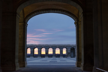 Horizontal view of a tunnel leading to a large courtyard lined with arches in the middle of a colorful sunset