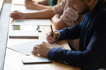 Close up of happy male job candidate put signature on paper contract after successful office interview, excited man worker or partner sign paperwork agreement, closing deal at team meeting