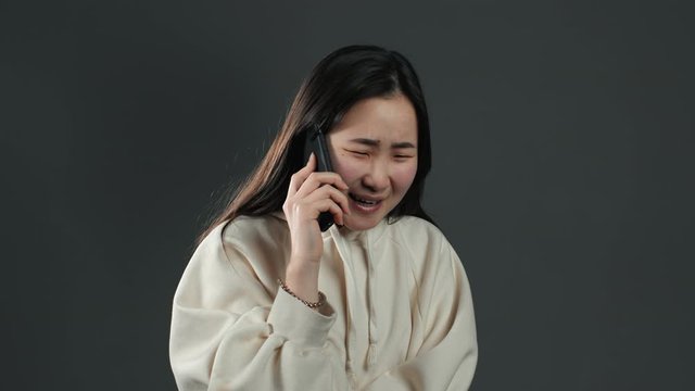 Pretty asian girl with long hair speaks on phone and sincerely surprised by news she heard, can't believe ears. Grey studio background