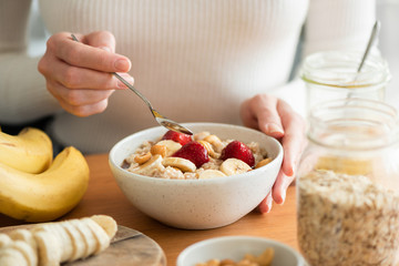 Woman eating oatmeal porridge with banana, strawberries and nuts. Healthy breakfast at the sunny morning kitchen table - obrazy, fototapety, plakaty