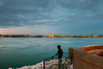 a woman in a jacket with a backpack looks at the Neva river on which ice floats on a winter evening