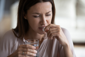 Head shot close up unhealthy young woman taking painkiller, suffering from strong headache. Unhappy frustrated lady holding glass of pure water and antidepressant medicine, healthcare concept.
