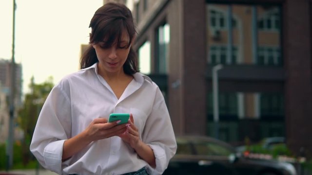 successful businesswoman holding mobile typing message walking on the street in city slow motion on the background modern architecture