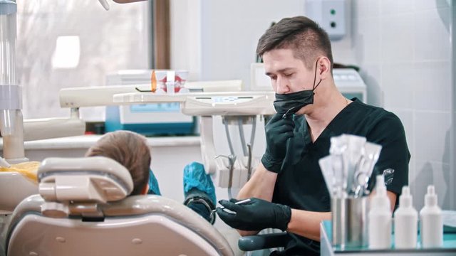 Young Man Dentist Putting On A Face Mask Before Checking The Mouth Of A Boy