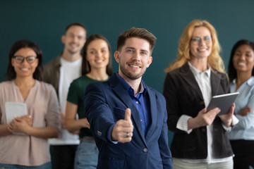 Confident millennial male team leader posing in office with diverse colleagues show thumb up give recommendation, successful businessman recommend good company business service, leadership concept