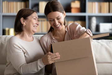 Happy older mature mommy sitting on couch with smiling grownup child, unpacking parcel together at home. Excited different female generations clients unboxing ordered goods from internet store.