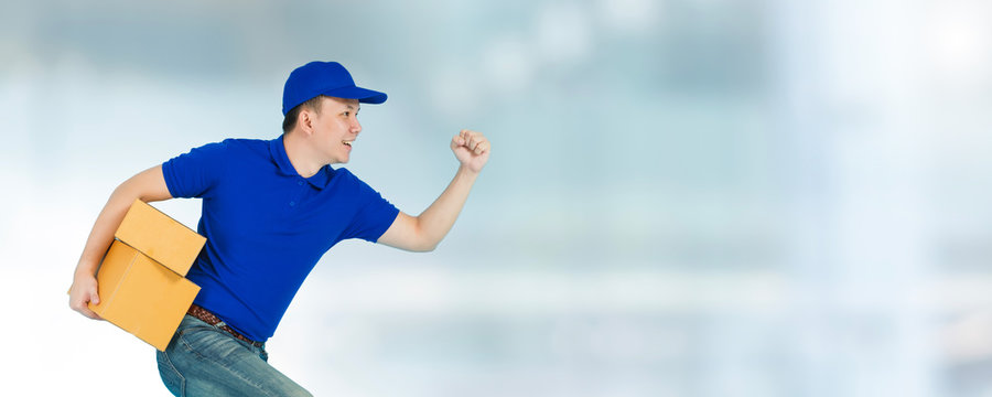 Asian happy delivery man wearing a blue shirt running and carrying paper parcel boxes isolated on blur window of shopping mall background with copy space.Concept of Postal delivery service.