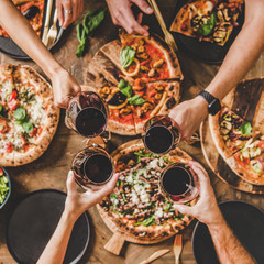 Family or friends having pizza party dinner. Flat-lay of people clinking glasses with red wine over rustic wooden table with various kinds of Italian pizza, top view, square crop. Fast food lunch