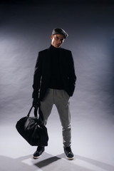 Portrait of a young man with sharp jawline wearing a newsboy cap, holding a man bag while posing in studio on a white background.