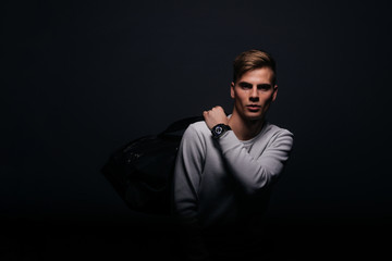 Fototapeta premium Informally ( casual ) dressed blonde young man with sharp jawline in his 20's posing in a studio in front of a black background while wearing a white sweater and holding a black man bag.