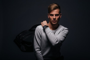 Obraz premium Informally ( casual ) dressed blonde young man with sharp jawline in his 20's posing in a studio in front of a black background while wearing a white sweater and holding a black man bag.