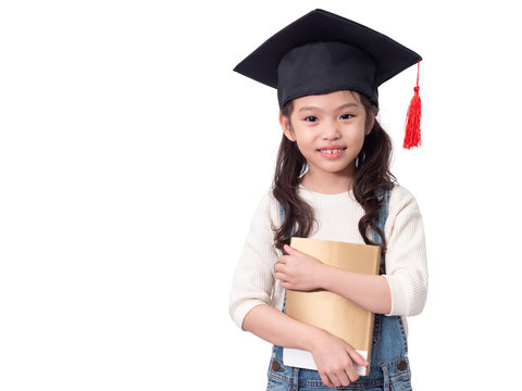 Asian Little Cute Girl 6 Years Old Wearing Graduation Hat And Holding A Book On Hands. Preschool Lovely Kid With A Book. 