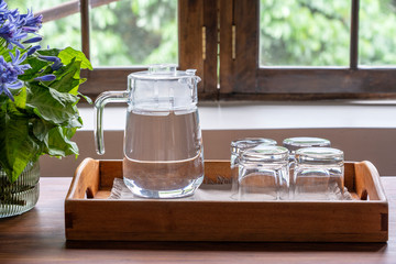 A carafe of water and four empty glasses on a wooden tray next to the window, closeup