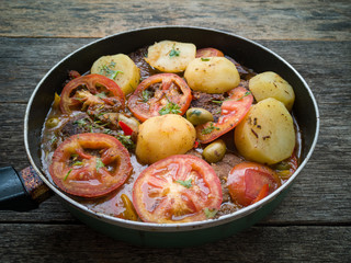 Meat with broth and vegetables in frying pan isolated on rustic wooden background.