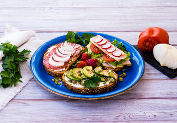 Three fresh sandwiches for a light and healthy eating - salami or roasted turkey or avocado and cheese - on blue plate. White wood background