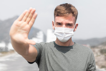 Guy, young man in protective sterile medical mask on his face looking at camera outdoors, on asian street show palm, hand, stop no sign. Air pollution, virus, Chinese pandemic coronavirus concept.