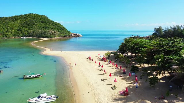 People sunbathe on white sandy beach with sunbeds and umbrellas near palm trees of vacations resort in front of turquoise lagoon in Koh phangan, Thailand