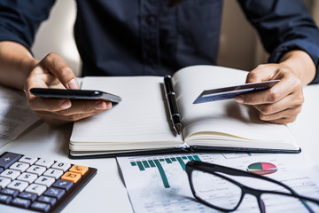 Young woman holding smartphone and calculating credit card expenses at home