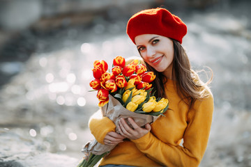 Beautiful young girl dressed in a red beret and orange sweater, holds a bouquet of red and yellow tulips in her hands and smiles. Portrait of a cute beautiful woman, copy space.