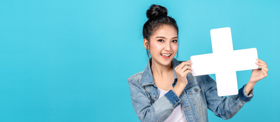 Banner of Happy asian woman smiling and holding plus or add sign on copy space blue background. Cute asia girl smiling wearing casual jeans shirt and showing join sign for increse and more concept - obrazy, fototapety, plakaty