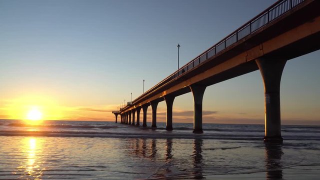 Tracking shot of sunrise above the sea in New Brighton, Christchurch of New Zealand; Waves under the bridge beating on the beach