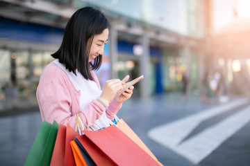 Happy Asian pretty girl holding shopping bags while using smartphone background shopping mall concept.