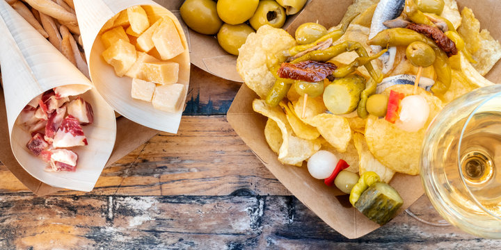 Tapas and pinchos overhead panoramic shot. Ham, cheese, olives, gildas with peppers and potato chips, with white wine, on a wooden table in an outdoors cafe