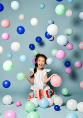 Fototapeta na wymiar Happy asian kid girl in stylish striped maxi skirt has fun sitting among plenty of colorful air balloons catching