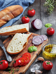 Fresh baguette sliced into pieces, tomatoes, vegetables and olive oil, ingredients for making a vegan sandwich, close-up. Dark wooden background.
