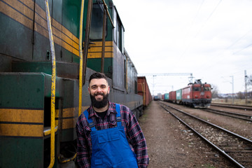 Portrait of engine train driver standing by locomotive at train station ready for departure. In background railroad and cargo containers.