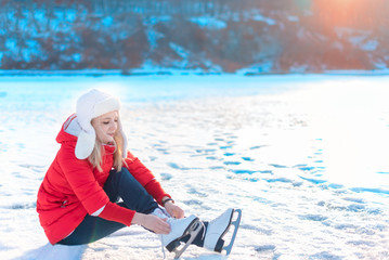 Young beautiful woman in red coat sits on ice and puts on figure skates early in the morning. Winter outdoor activities concept. Leisure and lifestyle. Background with copy space.