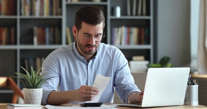 Focused businessman accountant doing calculation for online financial report at workplace. Serious man using calculator paying bill online holding paper sitting at home office desk. Accounting concept