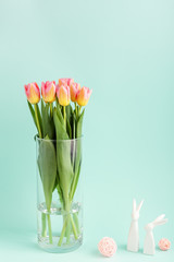 Pink and yellow tulip flowers in vase with easter bunnies on light aqua. Spring concept