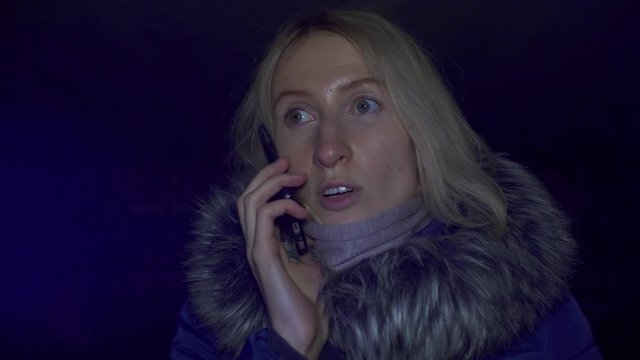 Portrait of shocked scared young woman. She is lit by police flashing lights and is talking on phone. woman is scared, desperately wants something, covering her mouth with hands of problem.