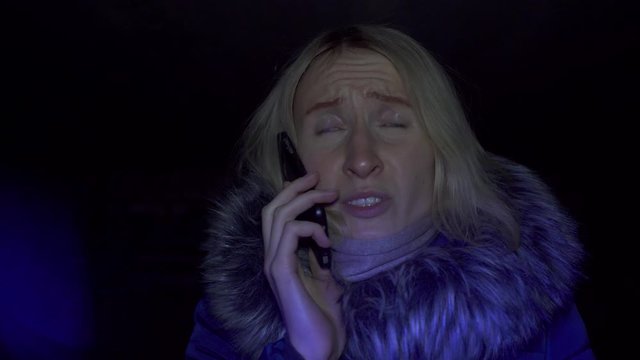 Portrait of shocked scared young woman. She is lit by police flashing lights and is talking on phone. woman is scared, desperately wants something, covering her mouth with hands of problem.