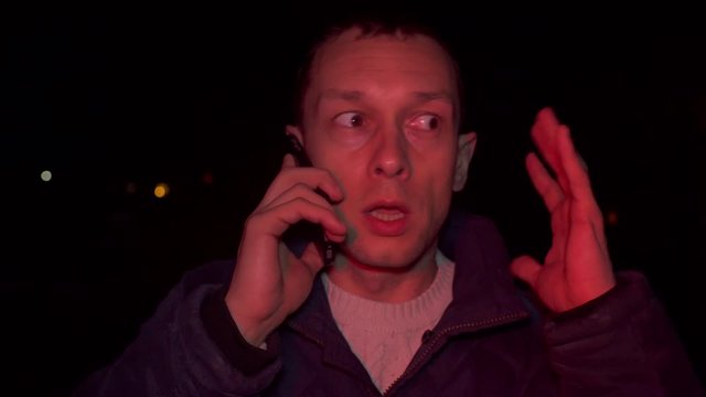 Portrait of shocked scared young man. he is lit by police flashing lights and is talking on phone. man is scared, desperately wants something, covering her mouth with hands of problem.