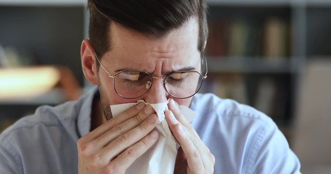 Ill young man having allergy symptoms blowing running stuffy nose in tissue. Unhealthy businessman suffering from sniffles, allergic rhinitis concept sitting at home or in office. Close up view