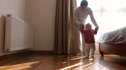 Slow motion of little asian baby girl holding her mother's hand learning to walking on the floor at home with afternoon sunlight coming from window 