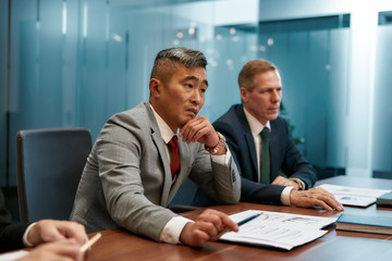 Fototapeta na wymiar Successful business partners. Portrait of caucasian and asian business people in formal wear sitting at office desk. Partners working together in the modern office